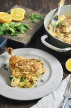 A chicken portion on a plate, surrounded by lemon slices and herbs. Cooking pan with more chicken
