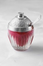 A bowl made of glass with a silver lid is filled with red lingonberry jam or sauce and sits on a