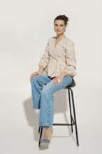 A woman sits on a black stool with a white background. She wears a light-colored blouse and blue