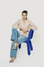 A woman sits on a blue chair with one leg crossed over the other. She wears a light top and denim