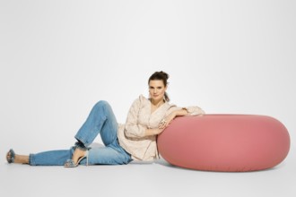 A woman in a light top and blue jeans is sitting next to a large pink cushion. She rests her arm on