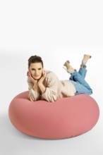 A woman lays on a big pink cushion in a studio. She is wearing a light blazer and jeans. Her hair