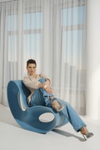 A woman is sitting on a blue chair. She is wearing casual clothes and is smiling. The room has a