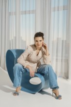 A woman sits on a blue chair with a unique design. She wears a light-colored top and blue jeans.