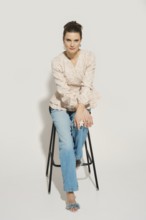 A model sits on a tall stool in a studio. She wears casual clothes including a light top and denim