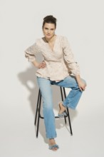 A model poses on a tall stool in a studio with a plain background. She wears a fashionable top and