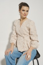 A woman sits on a black chair wearing a light-colored blazer and blue jeans. She has her hair