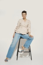 A woman poses on a black stool in a studio. She wears a light blazer with a floral design and blue