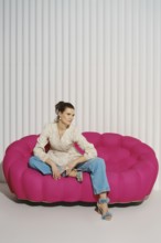 A woman is sitting on a large pink sofa in a modern indoor area. She wears a light-colored blazer