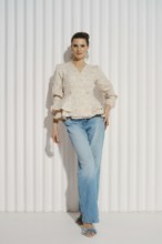 Relaxed woman stands with one hand on her hip. She wears a light blouse and blue jeans. The