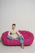 Woman sits with one leg tucked under her on a pink couch in a studio. She wears a light blazer and