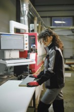 Woman wearing protective gloves and safety glasses runs a wooden piece into a machine for gluing