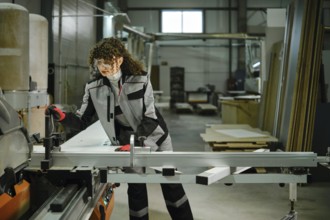 Woman is focused on using a large circular saw in a factory. The worker wears safety gear and