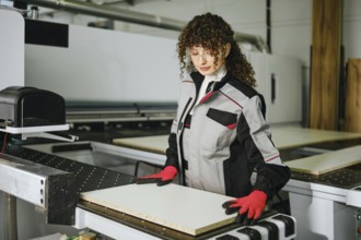 A person stands at a machine in a wood processing facility, wearing work gloves and a jacket. She