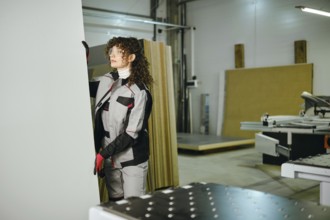 A worker prepares to cut a large sheet of wood in a workshop. The worker is dressed in protective