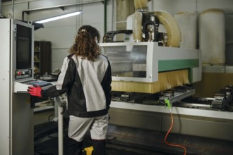 A woman uses a milling machine in a workshop. She stands with her back to the viewer and watches