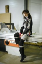 A woman is sitting on a large cutting machine in a workshop. She is dressed in work clothes and is