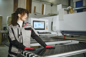 A woman stands beside wood cutting machine in carpentry workshop. She wears gloves and protective