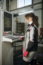 Woman stands at a control panel in a manufacturing facility. She wears a work uniform with gloves.