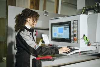 A woman using a computer to control cutting machine at furniture factory. She wears work clothing