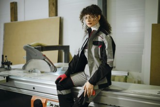 A woman sits on a machine in a workshop. She is wearing a work outfit and gloves. The workshop has