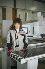 A woman stands in a workshop next to a wood shaping machine. She wears a work suit and gloves while
