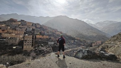 A hiker with backpack stands in front of a mountainous village in sunny weather, Atlas Mountains,