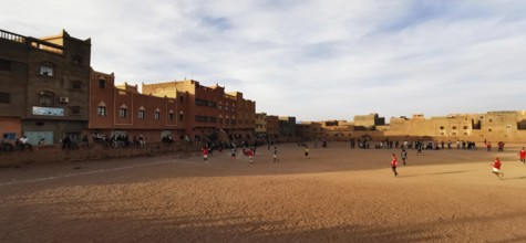Children playing soccer on a sandy playing field between buildings in cloudy sky, Morocco