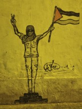 Graffiti of a masked person with Palestine flag showing the victory sign on yellow wall,