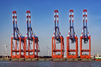 Set of red and blue cranes at the harbor, wind turbines and container warehouse in the background,
