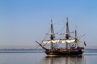 A traditional sailing ship with full sails on calm water under clear blue skies, an old historic