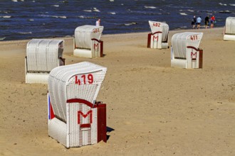 Several white beach chairs with red accents stand on the beach with walkers in the background,