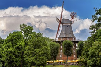 Historic windmill surrounded by trees under blue sky with white clouds and umbrellas, An old