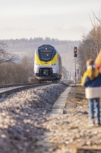 A yellow train travels on rails through a rural area with trees and people on the side of the road,