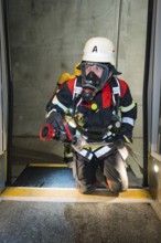 A firefighter wearing respiratory protection kneels in a tunnel at the threshold of a door, fire