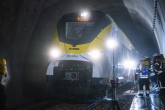 A train goes through an illuminated tunnel with firefighters in the background, fire department