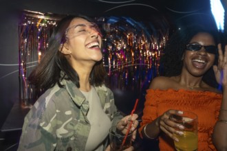 Two diverse young women enjoying a night out, laughing and dancing with drinks in hand amidst