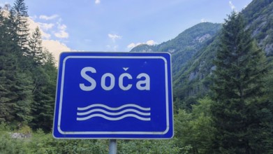 A blue road sign with the inscription 'Soca' in front of a mountainous forest landscape, Soca
