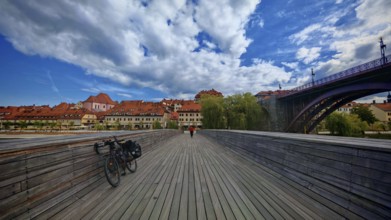 Wooden walkway with bicycle leading to a picturesque old town across the river under a blue sky