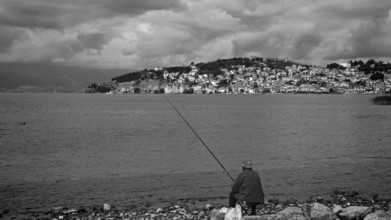 Black and white photo of a fisherman on a lake shore with a city in the background, Ohrid, North