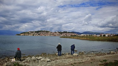 Fishermen on the shore of a lake with a view of a city in the background under cloudy sky, Ohrid,