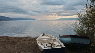 Two abandoned boats on the shore of a lake with mountains in the background under a cloudy sky,