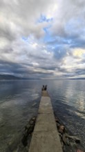 A long jetty leads into the calm sea under a cloudy sky that creates a peaceful atmosphere, Lake