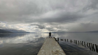 A man stands alone on a jetty that juts into a cloud-covered lake, Lake Ohrid, North Macedonia