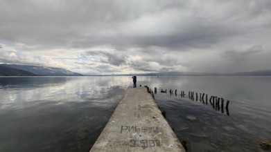 A man stands on a long jetty leading into a cloudy lake, Lake Ohrid, North Macedonia