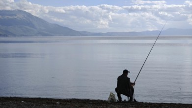 An angler sits on the shore of a peaceful lake, surrounded by mountains and a peaceful, natural