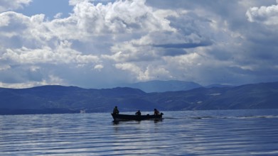 A small boat with silhouettes in a calm lake surrounded by mountains and dramatic cloud formations,