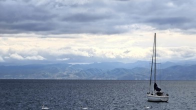 A sailboat on a calm sea with mountains in the background and cloudy sky, Lake Ohrid, North