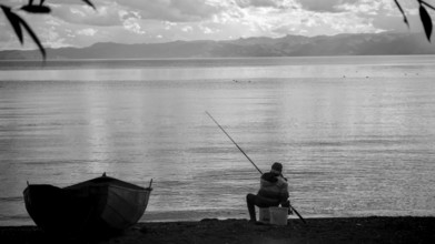 An fisherman sitting on the shore of a calm lake, with a small boat and a black and white