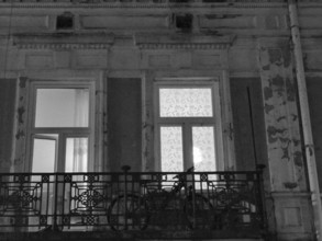 Worn façade at night with balcony on which a bicycle stands and illuminated windows in black and